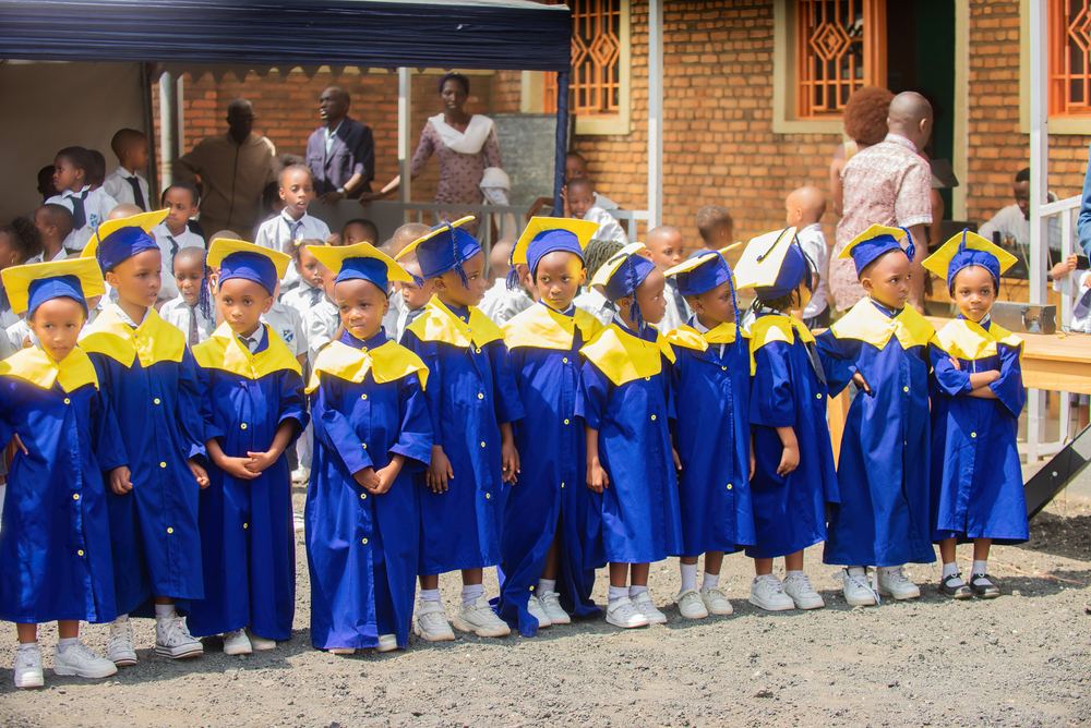 Nursery pupils in graduation gowns lined up in the school yard, with families behind them