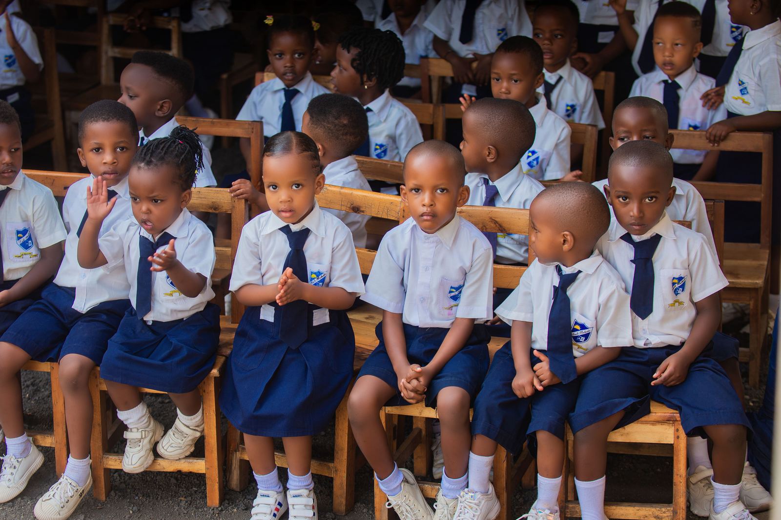 Nursery pupils in school uniform sitting on small wooden chairs at a school ceremony, one girl raising her hand
