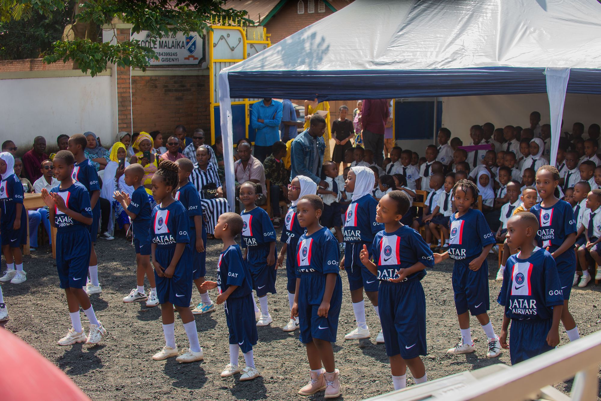 An event day in the school yard: pupils in football kit perform while families and classmates watch, beside the school gate and sign